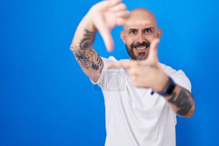 Téléchargez les photos : Homme hispanique avec des tatouages debout sur fond bleu souriant faisant cadre avec les mains et les doigts avec le visage heureux. concept de créativité et photographie. - en image libre de droit