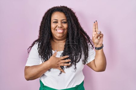 Foto de Mujer hispana de tamaño grande de pie sobre fondo rosa sonriendo jurando con la mano en el pecho y los dedos hacia arriba, haciendo un juramento de lealtad promesa - Imagen libre de derechos