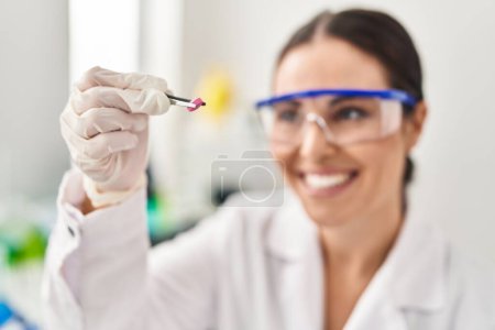 Foto de Joven hermosa mujer hispana científica sonriendo confiada sosteniendo píldora en laboratorio - Imagen libre de derechos