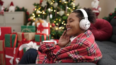 Téléchargez les photos : Femme afro-américaine écoutant de la musique boire du café assis près de l'arbre de Noël à la maison - en image libre de droit