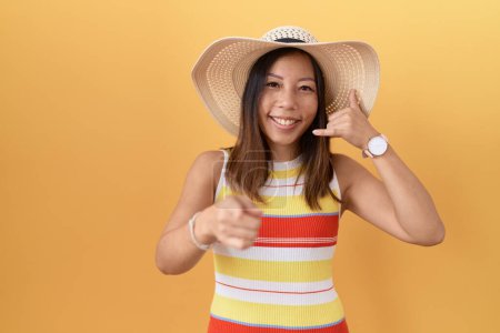 Foto de Mujer china de mediana edad con sombrero de verano sobre fondo amarillo sonriendo hablando por teléfono y señalándote. Llámame.. - Imagen libre de derechos