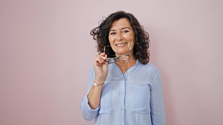 Téléchargez les photos : Moyen Age femme hispanique souriant confiant prendre des lunettes sur fond rose isolé - en image libre de droit