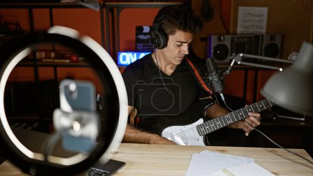 Foto de Joven músico hispano cantando una canción tocando un video de grabación de guitarra eléctrica por smartphone en un estudio de radio - Imagen libre de derechos