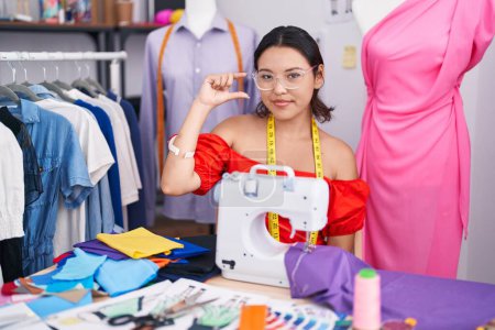 Photo for Hispanic young woman dressmaker designer using sewing machine smiling and confident gesturing with hand doing small size sign with fingers looking and the camera. measure concept. - Royalty Free Image