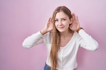 Photo for Young caucasian woman standing over pink background trying to hear both hands on ear gesture, curious for gossip. hearing problem, deaf - Royalty Free Image