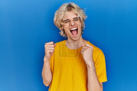 Téléchargez les photos : Jeune homme moderne debout sur fond bleu excité pour le succès avec les bras levés et les yeux fermés célébrant la victoire en souriant. concept gagnant. - en image libre de droit