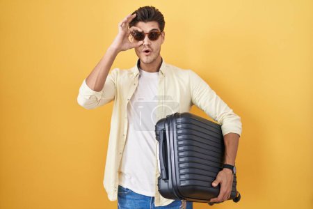 Foto de Un joven hispano sosteniendo una maleta de vacaciones de verano haciendo un buen gesto conmocionado con la cara sorprendida, los ojos mirando a través de los dedos. expresión incrédula. - Imagen libre de derechos