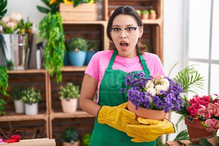 Téléchargez les photos : Jeune femme hispanique travaillant dans un magasin de fleuristes tenant l'usine effrayée et choquée par l'expression surprise et stupéfaite, la peur et le visage excité. - en image libre de droit