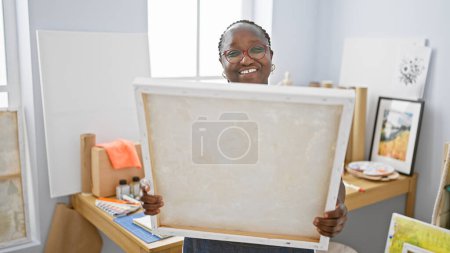Téléchargez les photos : Femme artiste afro-américaine confiante souriant, appréciant son passe-temps, tenant pinceau et peinture sur toile tout en regardant ses œuvres en studio - en image libre de droit