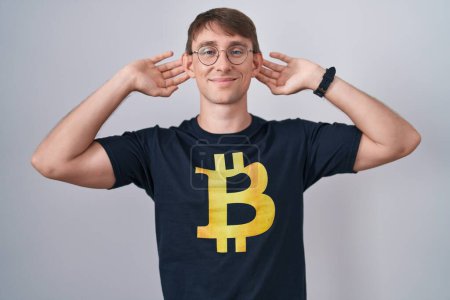 Foto de Hombre rubio caucásico con camiseta bitcoin sonriendo tirando de las orejas con los dedos, gesto divertido. problema de audición - Imagen libre de derechos