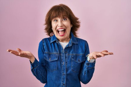 Foto de Mujer de mediana edad de pie sobre fondo rosa celebrando loco y sorprendido por el éxito con los brazos levantados y los ojos abiertos gritando emocionado. concepto ganador - Imagen libre de derechos