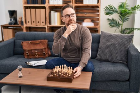 Photo for Middle age caucasian man playing chess sitting on the sofa serious face thinking about question with hand on chin, thoughtful about confusing idea - Royalty Free Image