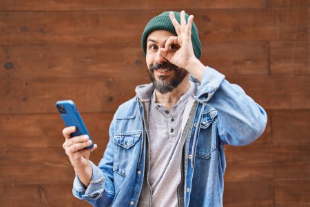 Téléchargez les photos : Jeune homme hispanique utilisant smartphone souriant heureux faire ok signe avec la main sur les yeux regardant à travers les doigts - en image libre de droit