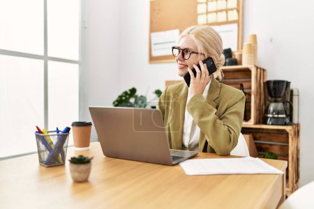 Téléchargez les photos : Jeune femme blonde travailleuse d'affaires utilisant un ordinateur portable parlant sur un smartphone au bureau - en image libre de droit