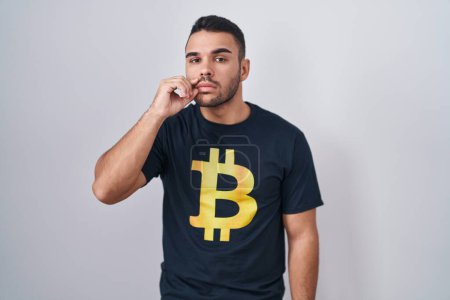 Téléchargez les photos : Jeune homme hispanique portant Bitcoin t-shirt bouche et lèvres fermées comme zip avec les doigts. secret et silencieux, tabou parlant - en image libre de droit