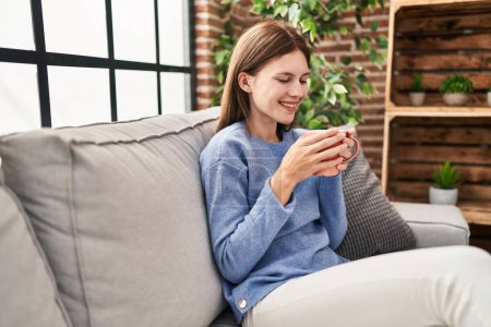 Téléchargez les photos : Jeune femme blonde buvant du café assis sur le canapé à la maison - en image libre de droit