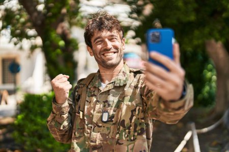 Photo for Hispanic young man wearing camouflage army uniform doing video call screaming proud, celebrating victory and success very excited with raised arm - Royalty Free Image