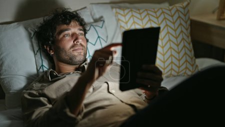 Téléchargez les photos : Jeune homme hispanique utilisant pavé tactile couché sur le lit dans la chambre - en image libre de droit