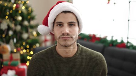 Photo for Young hispanic man sitting on sofa by christmas tree with relaxed expression at home - Royalty Free Image