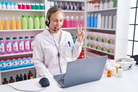 Téléchargez les photos : Jeune femme caucasienne travaillant à la pharmacie pharmacie en utilisant un ordinateur portable souriant avec un clin d'oeil heureux au visage à la caméra faisant signe de victoire. numéro deux. - en image libre de droit