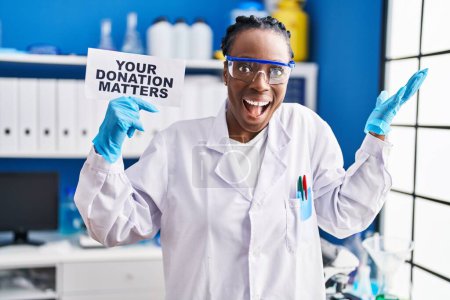Téléchargez les photos : Belle femme noire travaillant au laboratoire scientifique tenant votre bannière de questions de don célébrant la victoire avec sourire heureux et expression gagnante avec les mains levées - en image libre de droit