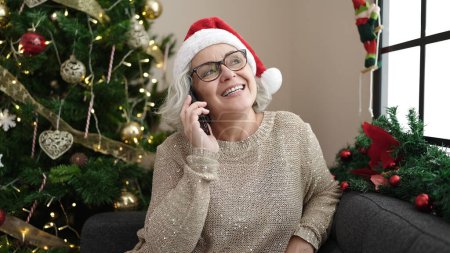 Photo for Middle age woman with grey hair talking on smartphone sitting on sofa by christmas tree at home - Royalty Free Image
