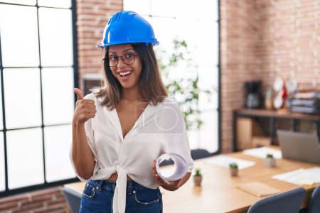 Photo for Hispanic young woman wearing architect hardhat at office smiling happy and positive, thumb up doing excellent and approval sign - Royalty Free Image