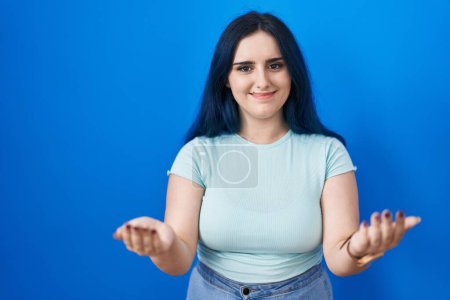 Foto de Joven chica moderna con el pelo azul de pie sobre fondo azul sonriente alegre ofreciendo las manos dando asistencia y aceptación. - Imagen libre de derechos