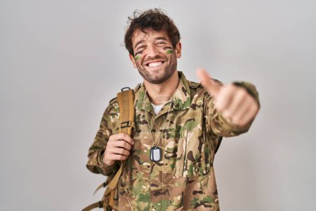 Foto de Joven hispano vistiendo uniforme camuflaje del ejército aprobando hacer gesto positivo con la mano, pulgares arriba sonriendo y feliz por el éxito. gesto ganador. - Imagen libre de derechos