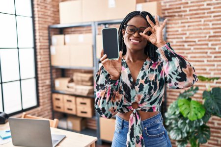 Téléchargez les photos : Femme africaine avec des tresses travaillant à la petite entreprise commerce électronique montrant écran smartphone souriant heureux faire ok signe avec la main sur les yeux en regardant à travers les doigts - en image libre de droit