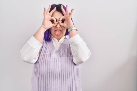 Téléchargez les photos : Plus taille femme avec des cheveux violets debout sur fond blanc faisant un geste correct comme des jumelles qui sortent la langue, les yeux regardant à travers les doigts. expression folle. - en image libre de droit