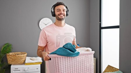 Photo for Young hispanic man listening to music holding basket with clothes at laundry room - Royalty Free Image