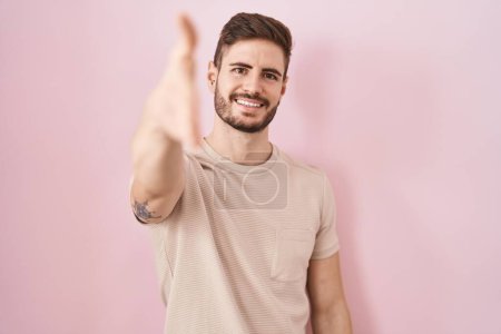 Foto de Hombre hispano con barba de pie sobre fondo rosa sonriente amable ofreciendo apretón de manos como saludo y bienvenida. negocios exitosos. - Imagen libre de derechos