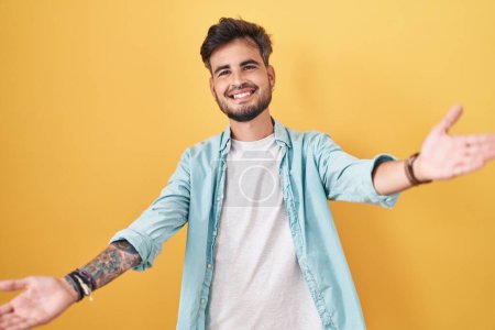 Téléchargez les photos : Jeune homme hispanique avec des tatouages debout sur fond jaune regardant la caméra souriant à bras ouverts pour un câlin. expression joyeuse embrassant le bonheur. - en image libre de droit