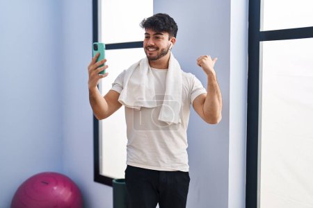 Foto de Hombre hispano con barba haciendo videollamada con smartphone en el gimnasio apuntando con el pulgar hacia un lado sonriendo feliz con la boca abierta - Imagen libre de derechos