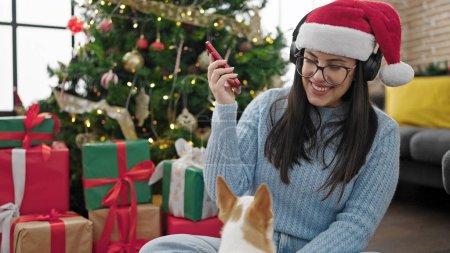 Photo for Young hispanic woman with chihuahua dog listening to music wearing headphones and christmas hat at home - Royalty Free Image