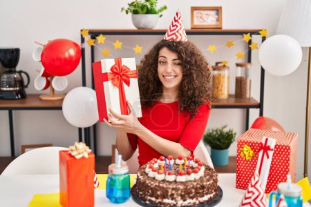 Téléchargez les photos : Femme hispanique aux cheveux bouclés célébrant l'anniversaire avec gâteau et clin d'oeil présent en regardant l'appareil photo avec expression sexy, visage joyeux et heureux. - en image libre de droit