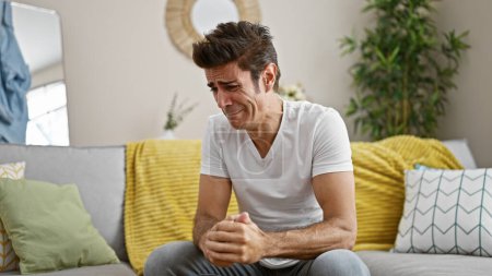 Téléchargez les photos : Jeune homme hispanique stressé assis sur le canapé pleurant à la maison - en image libre de droit