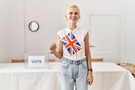 Téléchargez les photos : Jeune femme blonde tenant drapeau du royaume uni souriant au collège électoral - en image libre de droit