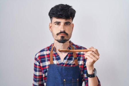 Photo for Young hispanic man with beard wearing apron tasting food holding wooden spoon thinking attitude and sober expression looking self confident - Royalty Free Image