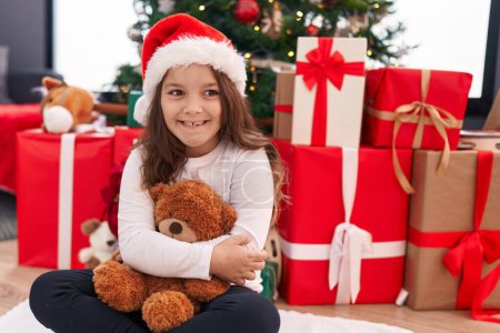 Photo for Adorable hispanic girl hugging teddy bear sitting on floor by christmas tree at home - Royalty Free Image