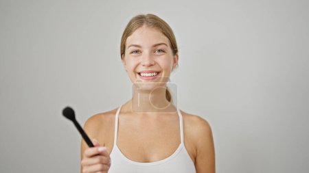 Téléchargez les photos : Jeune femme blonde souriante confiante tenant brosse de maquillage sur fond blanc isolé - en image libre de droit