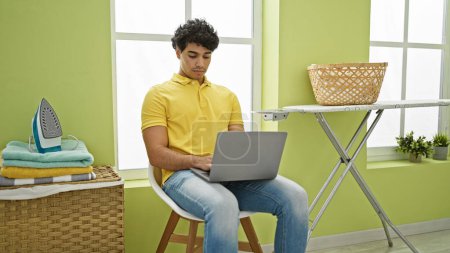 Photo for Young latin man using laptop waiting for washing machine at laundry room - Royalty Free Image