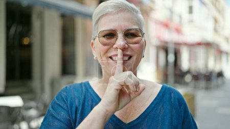Foto de Mujer de pelo gris de mediana edad con gafas de sol haciendo un gesto silencioso en la calle - Imagen libre de derechos