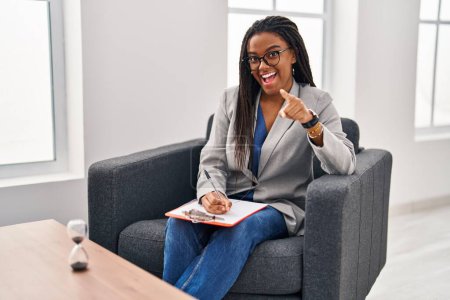 Téléchargez les photos : Jeune afro-américain avec des tresses travaillant au bureau de consultation vous pointant du doigt et la caméra, souriant positif et joyeux - en image libre de droit