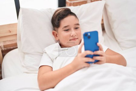 Foto de Adorable niño hispano usando smartphone acostado en la cama en el dormitorio - Imagen libre de derechos