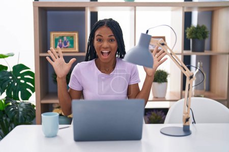 Foto de Mujer afroamericana con trenzas usando laptop en casa celebrando la victoria con sonrisa feliz y expresión ganadora con las manos levantadas - Imagen libre de derechos