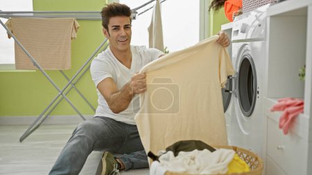 Téléchargez les photos : Jeune homme hispanique laver des vêtements tenant t-shirt souriant à la buanderie - en image libre de droit