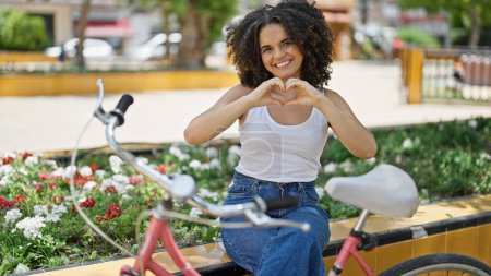 Photo for Young beautiful hispanic woman biker doing heart gesture sitting on bench at park - Royalty Free Image