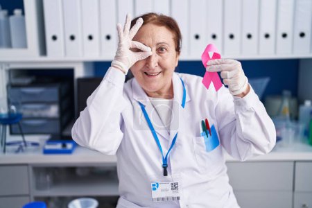 Foto de Mujer mayor con gafas trabajando en laboratorio científico sosteniendo cinta rosa sonriendo feliz haciendo signo de ok con la mano en el ojo mirando a través de los dedos - Imagen libre de derechos